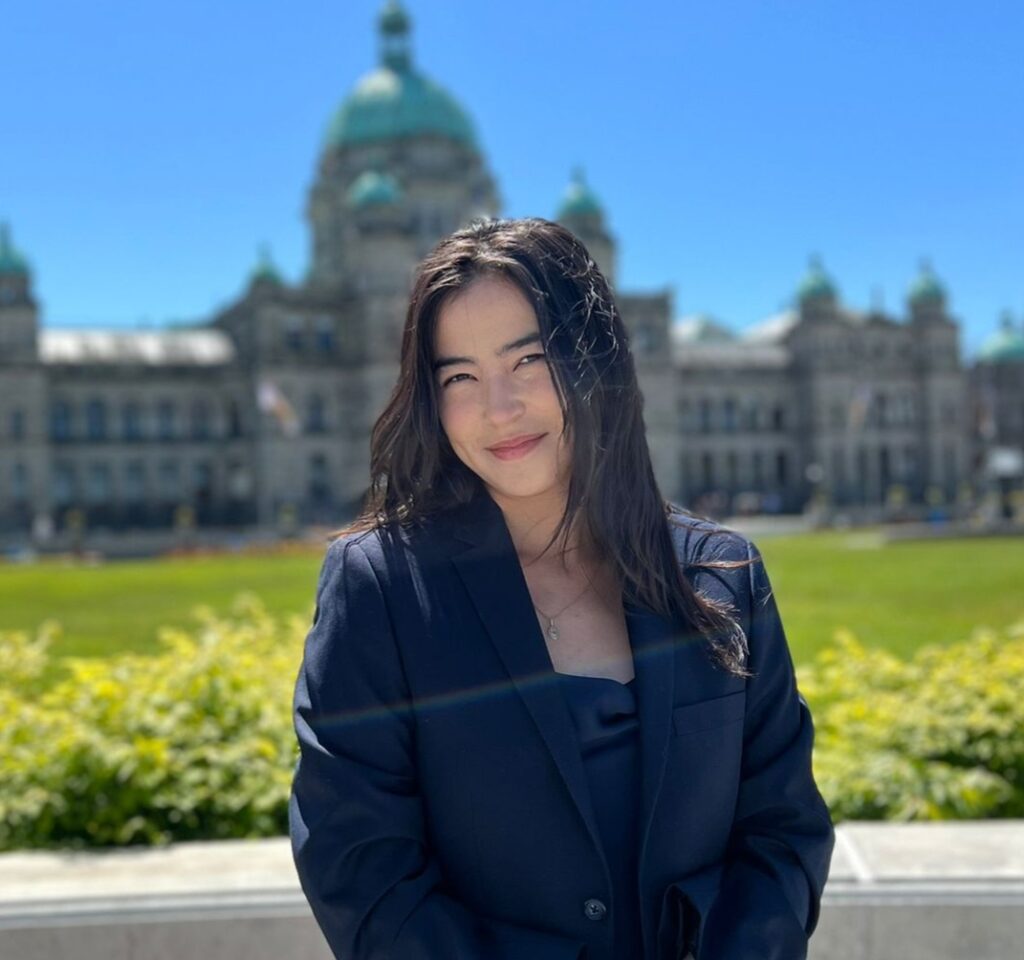 Emily Burlie Headshot A headshot of Emily Burlie, Speech-Language Pathologist at Granville Island Speech. She stands in front of a green field with a large building in the background.