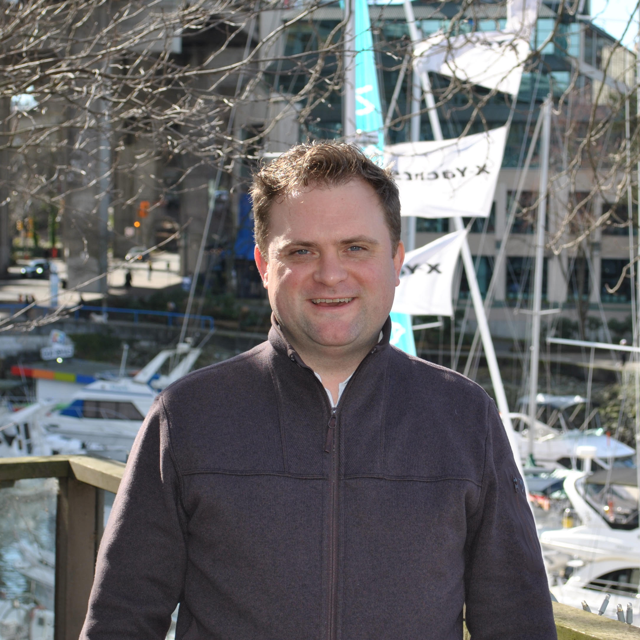 A headshot photo of Jamie Hack. He stands outside of the Granville Island Clinic with boats and the pier in the background. 